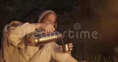 一个年轻的女人晚上坐在火对面的森林里，从<strong>热水</strong>瓶里倒茶<strong>喝</strong>。