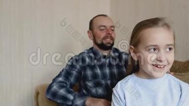 爸爸坐在沙发上梳女儿`头发。 爸爸和女儿花时间在一起。