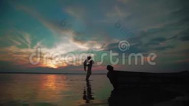 慢动作。 一个女人把孩子从湖边抱起来，在日落时和她一起在湖边快乐地转过身。 剪影式