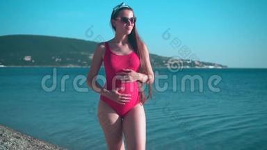 一个穿着红色一件泳衣和眼镜的怀孕女孩站在海边抚摸肚子。
