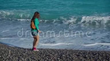 快乐的年轻女子，穿着绿色夹克、牛仔短裤和运动鞋，背着背包在温暖的阳光下沿着大海散步