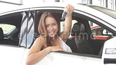 快乐的白种人年轻女司机坐在白色的车上，在汽车陈列室里展示汽车钥匙