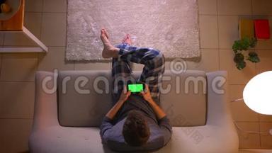 特写镜头：一个年轻人坐在室内沙发上用绿色屏幕在手机上玩电子游戏。