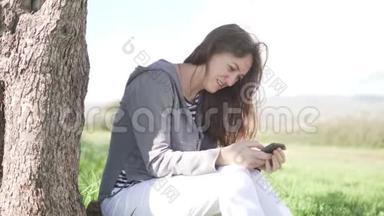 一个年轻的女人坐在树下的绿草上看着电话