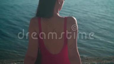 一个穿着红色一件泳衣的漂亮女孩坐在夕阳下的海滨。 那个女孩坐在相框里。