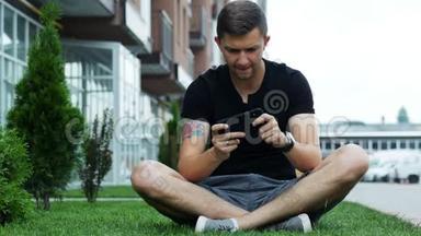 快乐男人在智能手机上玩手机游戏，坐在房子附近的草地上。