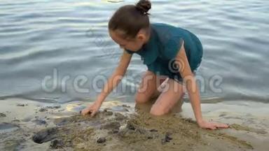 一个孩子，一个年轻的女孩，在日落时在河岸上建造一座沙堡。 她坐在水边`耙
