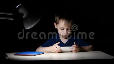 一个孩子晚上在家看卡通片，他用智能手机坐在桌子旁，被一盏灯照亮