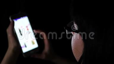 一个戴着眼镜的深色头发的女孩在晚上用手机特写，通过一家<strong>网上</strong>商店看<strong>网上</strong>购物