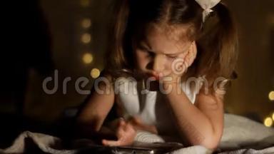 小女孩躺在床上，在黑暗中玩智能手机。 孩子的近脸被明亮的灯光照亮