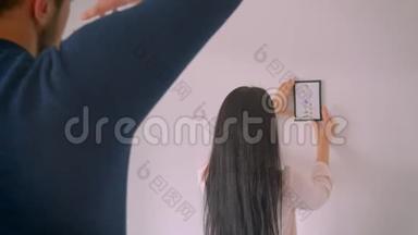 白种人的女孩正在选择白色墙壁上的相框图片的位置，而她的男朋友用手指做相框形状