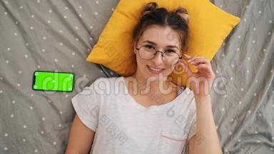 躺在床上的戴着眼镜的年轻迷人女孩的俯视图中，她兴高采烈地对着镜头笑着，用一个