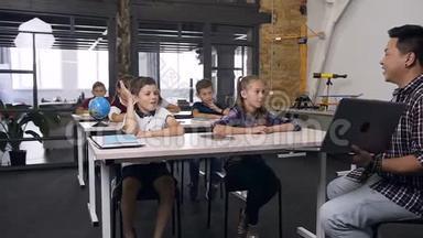 可爱的白种人男孩和同学坐在桌子后面，举手向韩国老师提问