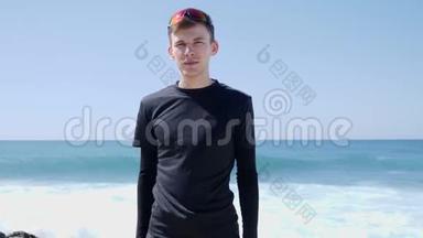 年轻的身材，英俊的<strong>黑衣</strong>人，竖起大拇指，对着站在海滩上的摄像机。 强烈的海浪在撞击Bea