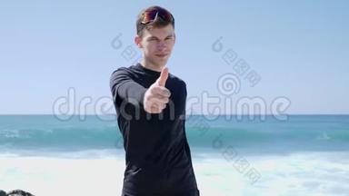 年轻的身材，英俊的黑衣人，竖起大拇指，对着站在海滩上的摄像机。 强烈的海浪在<strong>撞击</strong>Bea