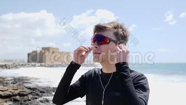 戴太阳镜的年轻运动员在训练前戴上耳机站在海滩上。 波浪正在撞击海岸线。