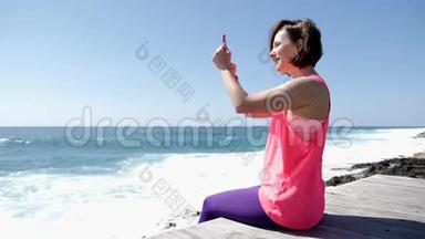 年轻美丽的白种人女人坐在岩石海滩上拿着电话，拍着海浪溅到岩石上的照片。