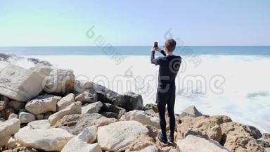 年轻的帅哥在一个阳光明媚的多风天气里，拍着海洋和海浪拍打着岩石海滩，<strong>溅起水花</strong>。 慢慢慢慢