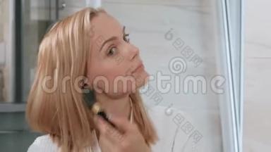 漂亮的中年金发女人穿着白色浴袍站在镜子旁边的浴室里。 化妆用的。