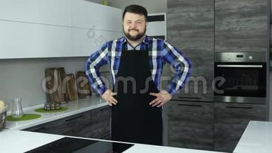 一个穿着围裙的胖子站在厨房的中央，双手放在一边微笑。 一个穿着围裙的胖子