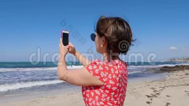 一个女人用手机站在海滩上拍照的特写镜头。 强风海浪背景上..