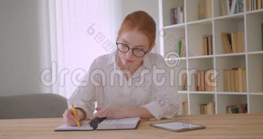 一位年轻漂亮的红头发女学生，戴着眼镜，用平板电脑写着一本笔记本，坐在