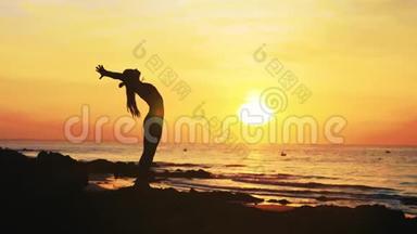 年轻女子的剪影正在日落时在海边做瑜伽练习。