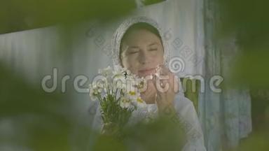 漂亮的老年妇女头上戴着白色披肩，在户外晾衣绳上撕下雏菊花瓣。 洗衣日