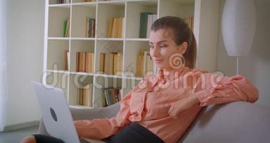 在笔记本电脑上工作的年轻漂亮女商人的特写镜头，她看着相机，兴高采烈地坐在电脑上