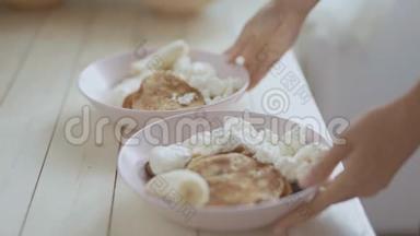 特写两个粉红色的碗，里面有煎饼、水果干酪和酸奶油。 女人从白色的木头上拿碗