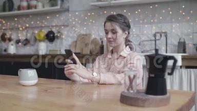 年轻的女人坐在家里的厨房桌子旁，用智能手机做她的小生意。 库存数据