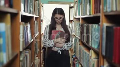 长着黑色长发的漂亮女孩直视镜头，在<strong>图书馆</strong>的<strong>书架</strong>上走着
