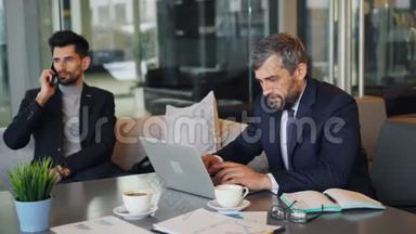 商人在咖啡馆里使用笔记本电脑，而合伙人则在谈论手机