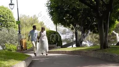 快乐的一对夫妇穿过绿色公园。 行动。 美丽的<strong>新婚</strong>夫妇在阳光下穿过公园。 年轻<strong>夫妻</strong>