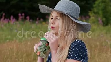 一个年轻漂亮的金发女郎戴着帽子，穿着裙子，坐在绿色的格子花格子上喝着罐装柠檬水