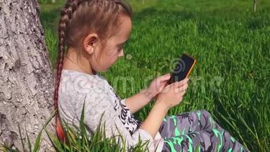 一个可爱的小女孩坐在树下的草地上，手里拿着电话。