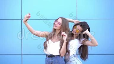 时尚肖像两个美丽的微笑和女人戴着太阳镜与智能手机靠在五颜六色的蓝色墙壁。 制作
