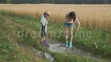 两个穿着靴子的快乐女孩姐妹在乡间小路上的雨水中嬉戏