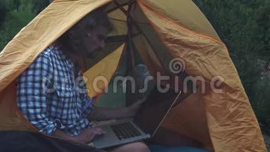 自由职业者使用笔记本电脑坐在森林的露营帐篷。 从事新启动项目的自由职业者