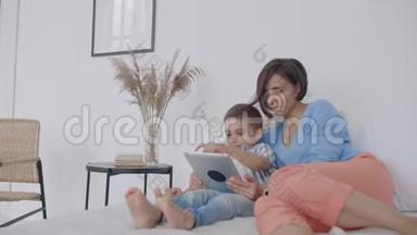 在白色的卧室里，妈妈和儿子看着平板电脑，笑了起来。 快乐的一家人在床上晨读一本书