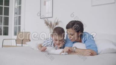 在白色的卧室里，妈妈和儿子看着平板电脑，笑了起来。 快乐的一家人在床上晨读一本书