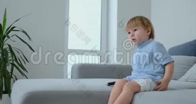一个2岁的男孩坐在沙发上看电视，手里拿着遥控器。 看动画片和电视