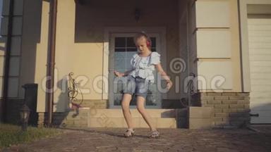 戴着耳机的可爱小女孩在傍晚的阳光下在房子附近旋转跳舞。闲暇少女