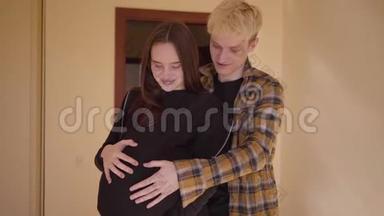 快乐的青少年夫妇站在家里在一起。 那个年轻人抚摸着怀孕女朋友的肚子。 <strong>未成年人</strong>