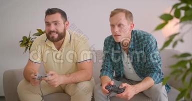 两位白种人成年男子在室内坐在沙发上玩电子<strong>游戏</strong>的特写镜头。 男子获胜