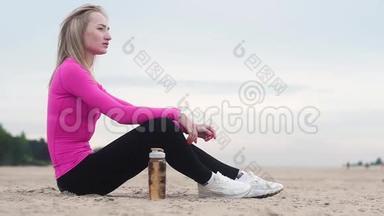 美丽的年轻运动女子在海滩上训练后休息。 从塑料瓶里喝水。