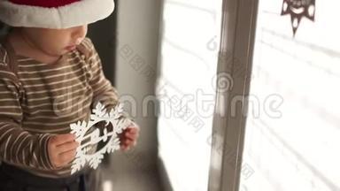 戴着圣诞帽的小宝宝在室内对着窗户玩圣诞装饰。 新年快乐，圣诞快乐
