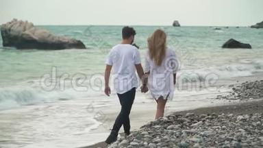 美丽的年轻夫妇在海滩上<strong>牵手</strong>，在石头上<strong>散步</strong>。 海浪飞溅。 令人惊叹的景色