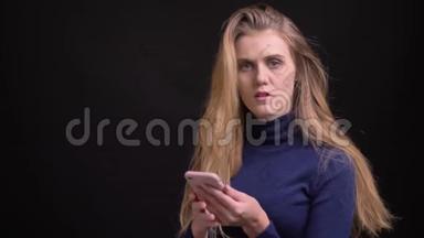 一位年轻的金发模特正在使用智能手机，她<strong>抬头</strong>看着镜头，在黑色背景下微笑着进入镜头。