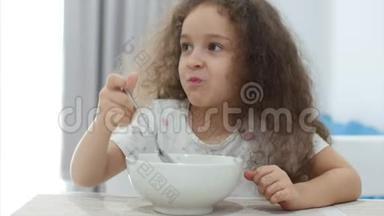有着卷发的<strong>可爱小</strong>女孩正坐在一张桌子旁，吃着自己的燕麦片，孩子愿意吃。 概念快乐